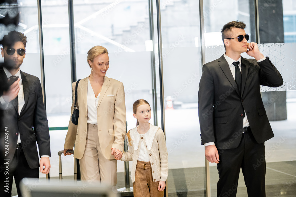 personal security and protection, two bodyguards in suits and sunglasses communicating while standing near hotel entrance, happy mother and child entering lobby, luxury lifestyle personal security and protection, two bodyguards in suits and sunglasses communicating while standing near hotel entrance, happy mother and child entering lobby, luxury lifestyle