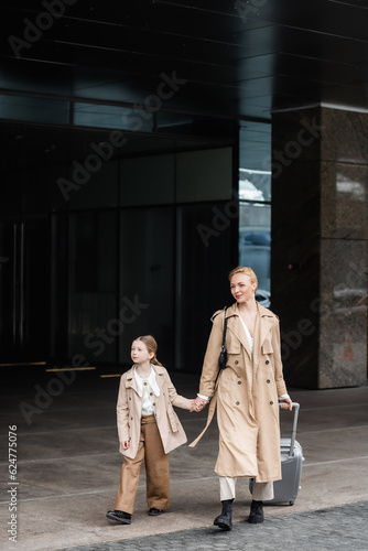 autumn fashion, mother daughter time, happy woman with luggage holding hand of preteen kid while walking out of hotel together, smart casual, beige trench coats, outerwear, trendy look