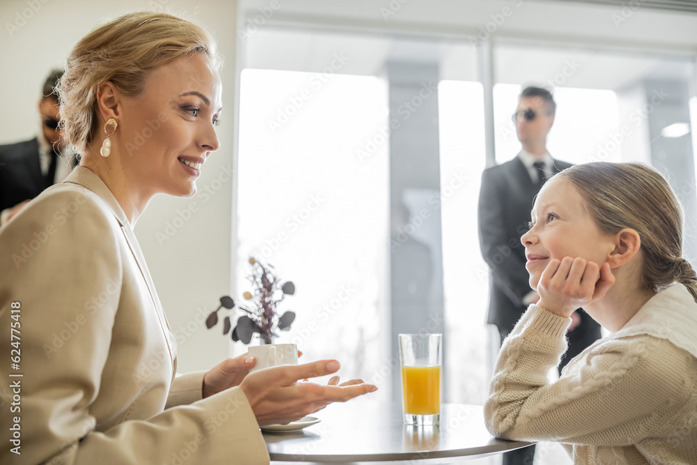 Fototapeta premium security service, private safety concept, cheerful blonde woman with cup of coffee talking with preteen daughter in cafe, bodyguards on blurred background, beverages and communication