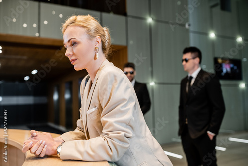 successful blonde woman in formal wear standing at reception desk, personal security service concept, two bodyguards in suits on blurred background, hotel industry, luxury travel, formal wear