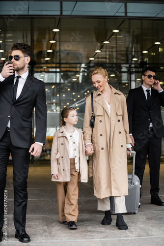 handsome bodyguards walking next to happy woman and preteen kid, entering hotel, private security, mother holding hands with daughter and wearing trench coats, safety and protection
