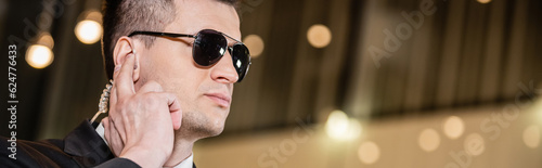 handsome bodyguard in sunglasses, handsome man in suit and tie touching earpiece in lobby of hotel, security, career, communication, vigilance, private safety, hotel staff, banner