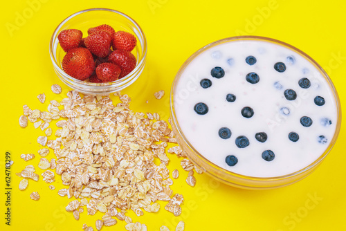 Glass bowl of yogurt with fresh blueberries, bowl with strawberries and a lot of scattered cereals on yellow background.