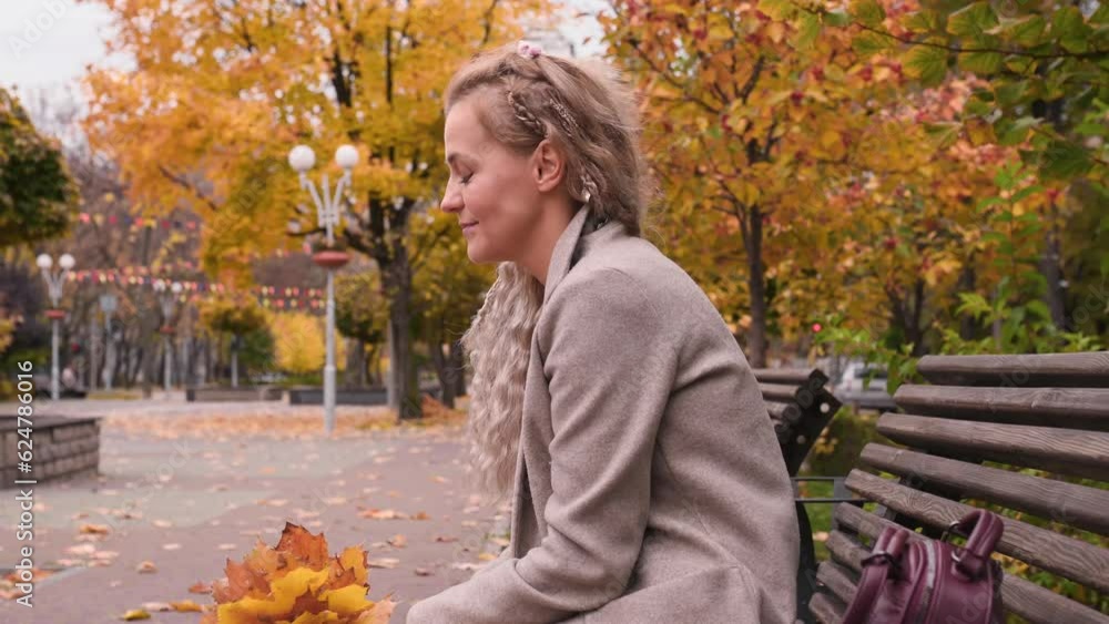 A beautiful mature blonde woman with pigtails in a trendy beige coat sits resting on a bench in a city park. Middle-aged woman enjoying autumn in the city A beautiful mature blonde woman with pigtails in a trendy beige coat sits resting on a bench in a city park. Middle-aged woman enjoying autumn in the city