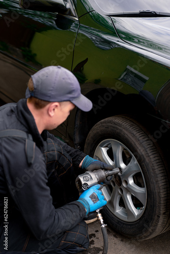 Auto mechanic in a cap changing a wheel from a black car using a drill in a tire shop on the street detailing car repairs