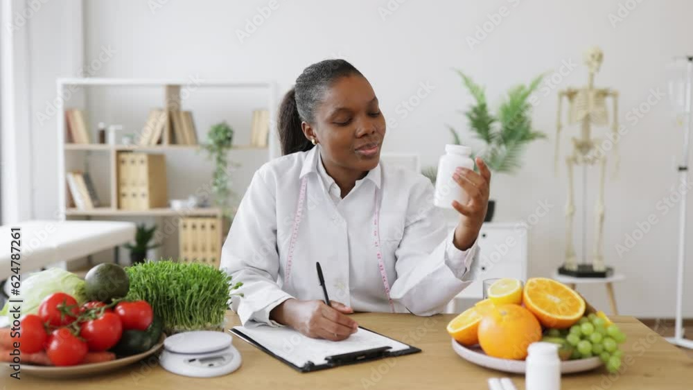 Confident multiethnic female with medicine container in hand recording new ideas of diet plan in workplace. Efficient nutritionist recommending meal modifications with food supplements in hospital. Confident multiethnic female with medicine container in hand recording new ideas of diet plan in workplace. Efficient nutritionist recommending meal modifications with food supplements in hospital.