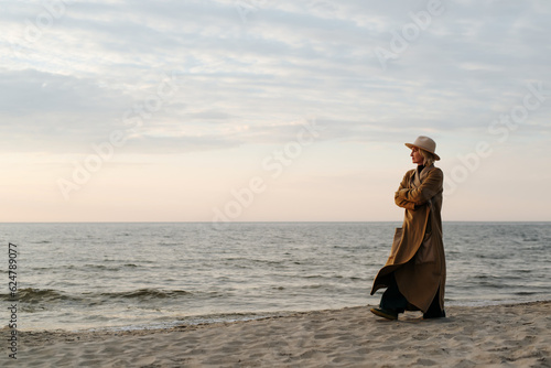 Thoughtful lonely stylish senior woman in brown coat and hat walking on beach in evening, copy space. Adult caucasian lady alone looking at sea