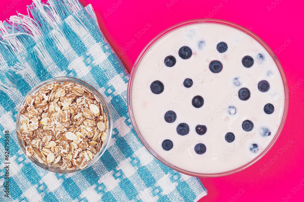 Glass bowl of yogurt with fresh blueberries and bowl with cereals on pink background. Top view. Glass bowl of yogurt with fresh blueberries and bowl with cereals on pink background. Top view.