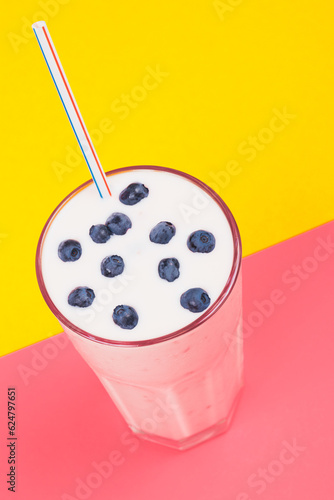 Glass of cold milkshake with fresh blueberries on top. Drink in a glass with a straw on a yellow and pink background. Multicolored background.