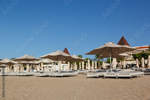 Row of sun loungers and umbrellas on the empty beach during sunrise awaits tourists. Blue sky background