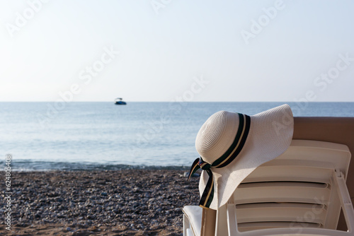 Straw women's hat on a back of a sun lounger. Vacations and accessories concept. Hat focus. Sea and sand background. Back view. 