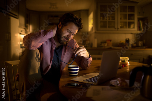 Young man going over his bills and finances late at night in the kitchen of his home and having neck pain photo