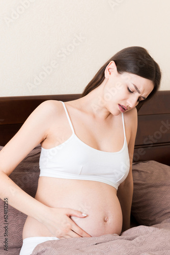 Young pretty pregnant woman, in pain, sitting on bed in bedroom, preparing to give a birth