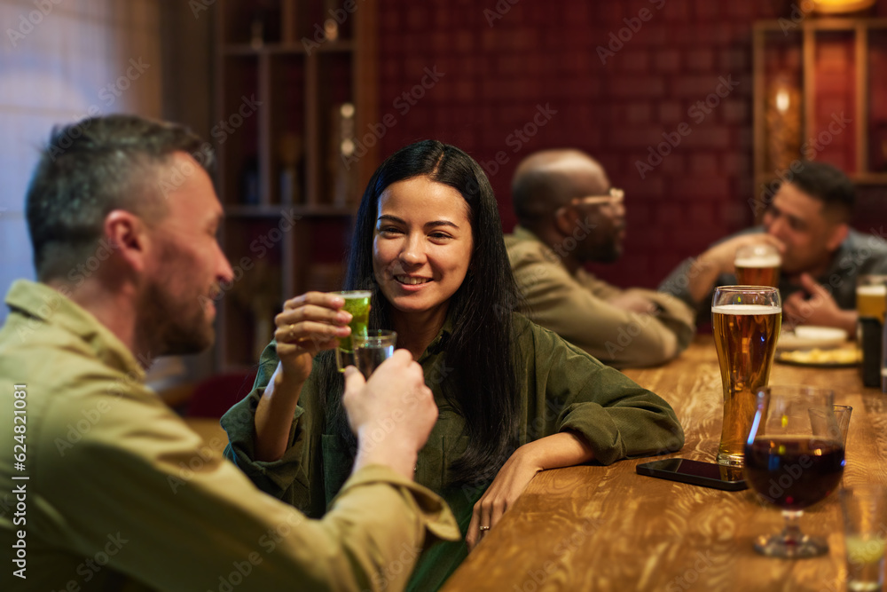 Happy young brunette woman with glass of aperitif toasting with her boyfriend while both sitting by bar counter and celebrating life event