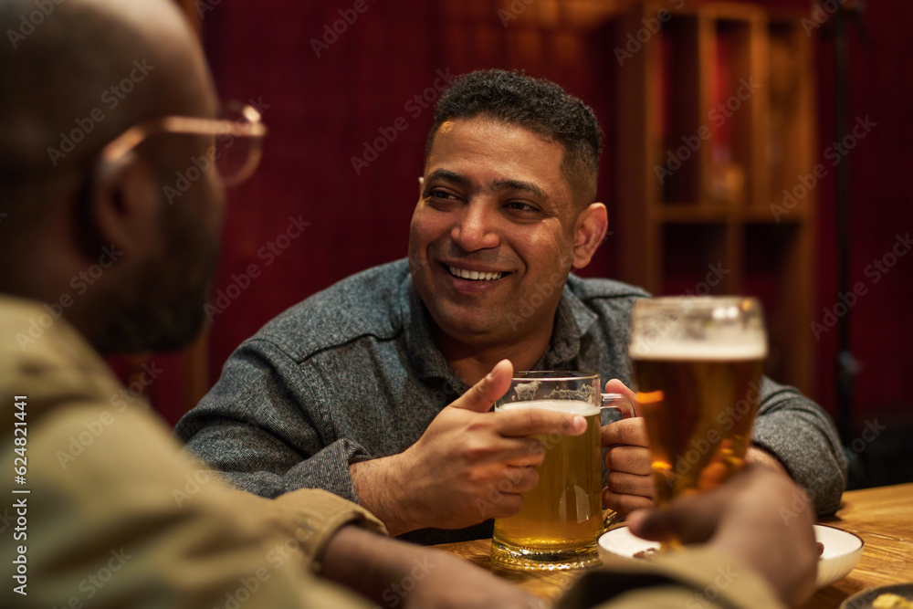 Focus on happy young man with big mug of lager beer looking at his African American buddy during chat while both sitting by bar counter