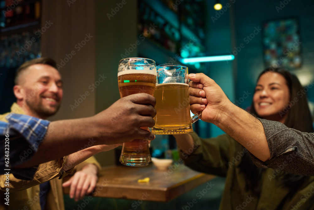 Focus on hands of intercultural buddies clinking with glasses and mugs of refreshing beer while sitting by table and bar counter and toasting Focus on hands of intercultural buddies clinking with glasses and mugs of refreshing beer while sitting by table and bar counter and toasting