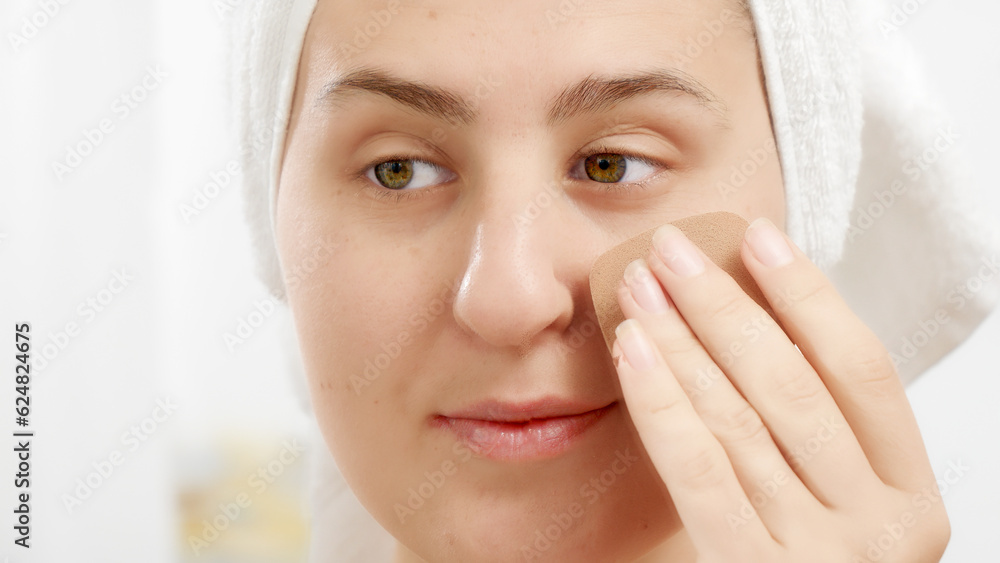 Closeup of young woman in bath towel applying cosmetics on face with soft sponge. Concept of beautiful female, makeup at home, skin care and domestic beauty industry