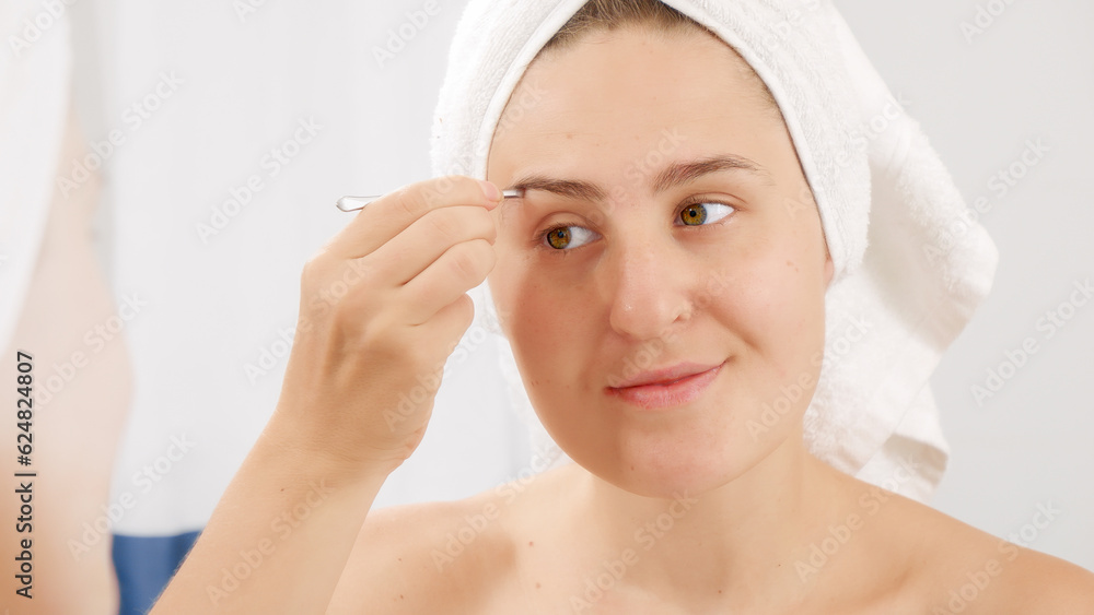 Young woman in bath towel using tweezers for plucking eyebrows. Concept of beautiful female, makeup at home, skin care and domestic beauty industry.
