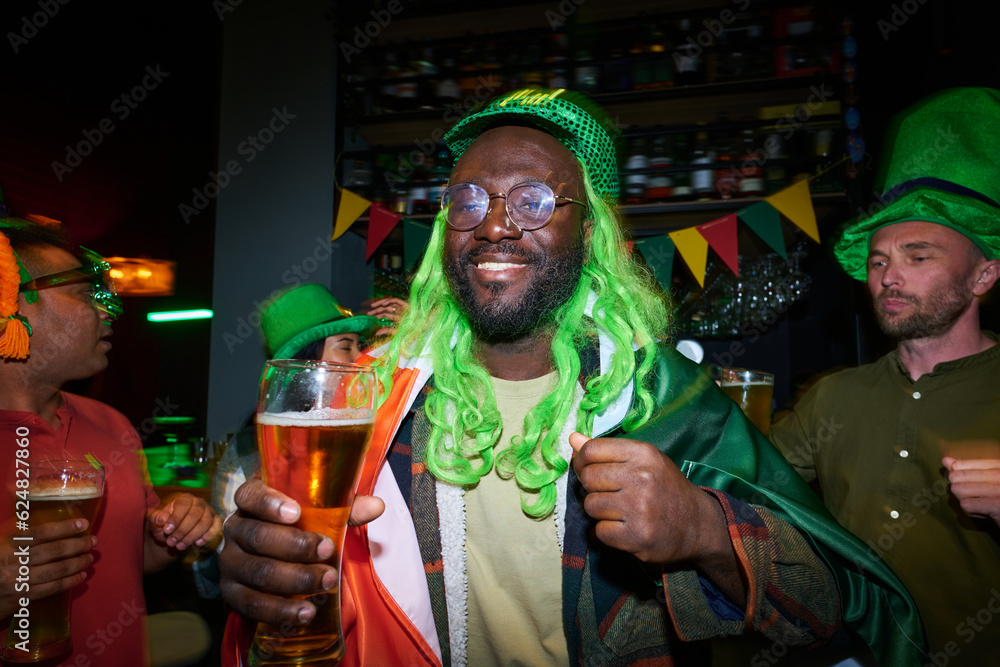 Happy young African American man in green cap and wig dancing at party in Irish pub against group of intercultural buddies with glasses of beer