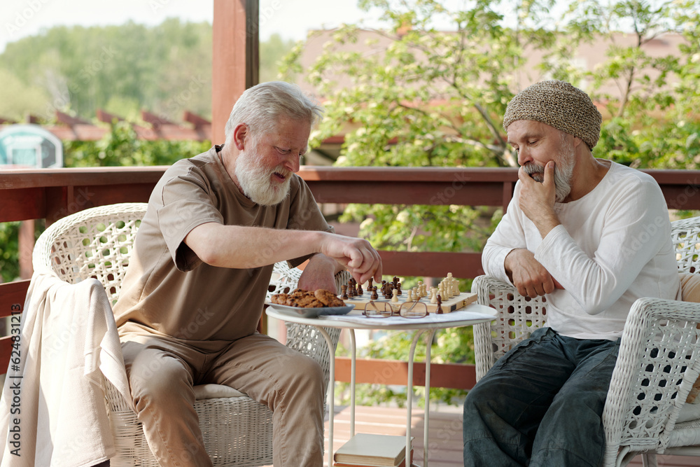 Two bearded senior men in casualwear playing chess by small round table after breakfast while sitting in armchairs on patio of retirement home Two bearded senior men in casualwear playing chess by small round table after breakfast while sitting in armchairs on patio of retirement home
