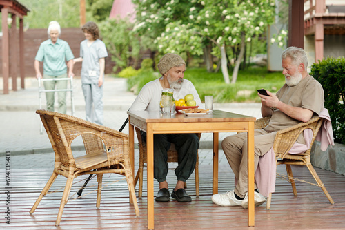 Two senior men sitting in comfortable wicker armchairs by wooden table on patio of retirement house while one of them scrolling in smartphone © pressmaster