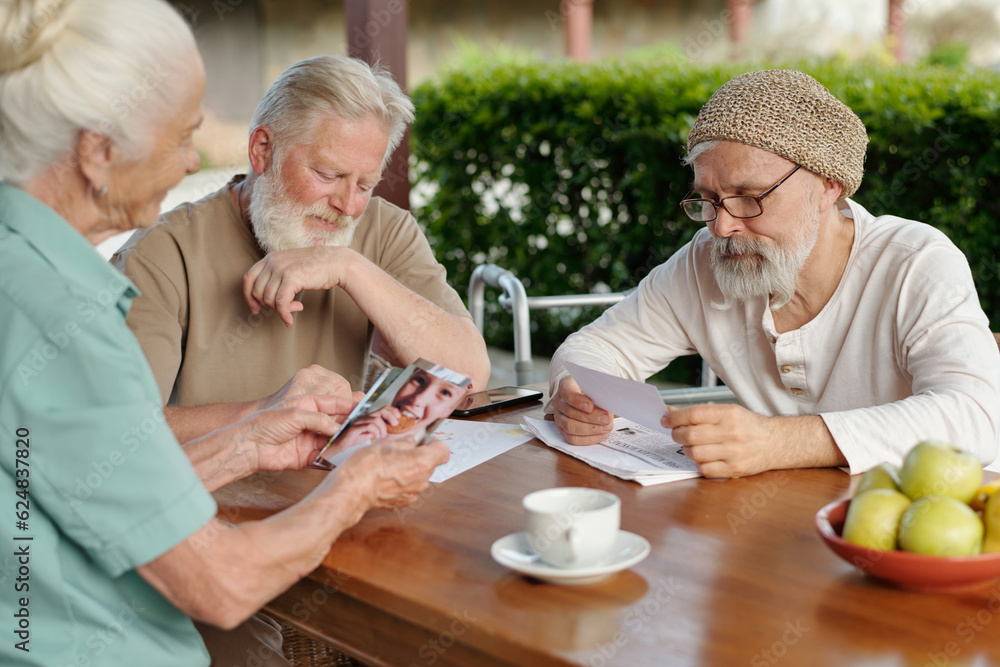 Two elderly bearded men and grey haired woman looking through photos of their family and grandchildren while sitting by table after breakfast Two elderly bearded men and grey haired woman looking through photos of their family and grandchildren while sitting by table after breakfast