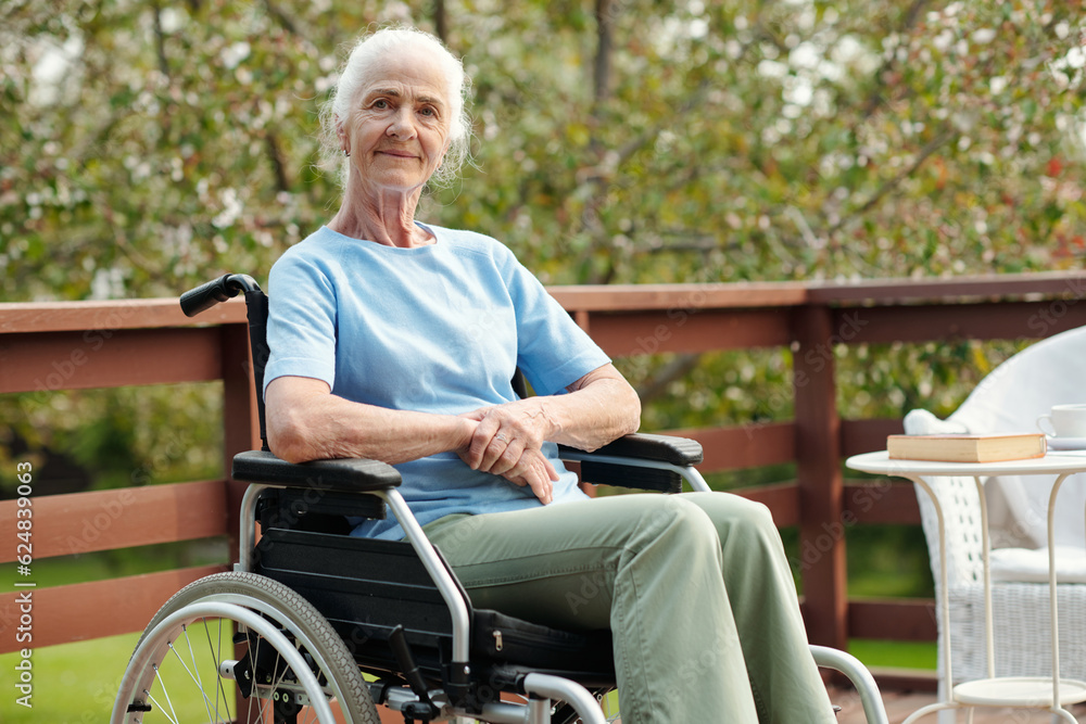 Elderly woman with white hair sitting in wheelchair in front of camera against garden and territory of retirement home and looking at you Elderly woman with white hair sitting in wheelchair in front of camera against garden and territory of retirement home and looking at you