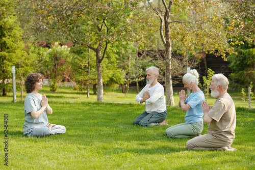 Fototapeta Naklejka Na Ścianę i Meble -  Side view of young female yoga instructor and group of senior patients of retirement home practicing meditation exercise on green lawn