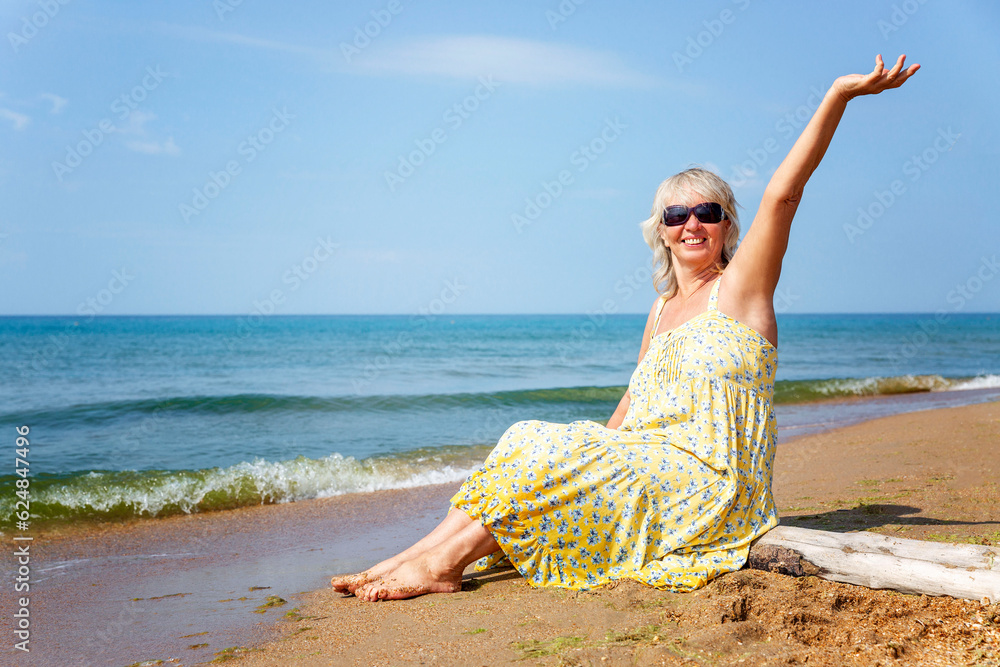 Laughing elderly woman in a yellow sundress and sunglasses sits on a sandy seashore. Happiness, positive, vacation at the resort and an active lifestyle. Laughing elderly woman in a yellow sundress and sunglasses sits on a sandy seashore. Happiness, positive, vacation at the resort and an active lifestyle.