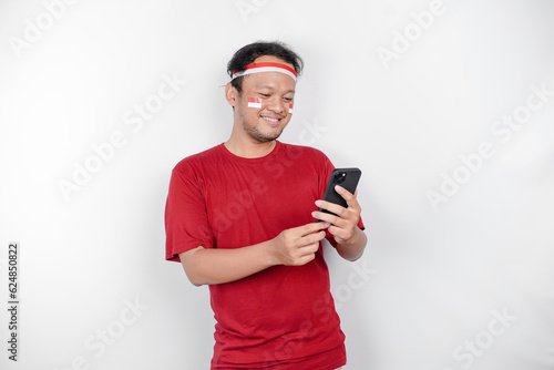 A portrait of a smiling Asian man wearing headband and holding his phone, isolated by white background. Indonesia's independence day concept © Reezky
