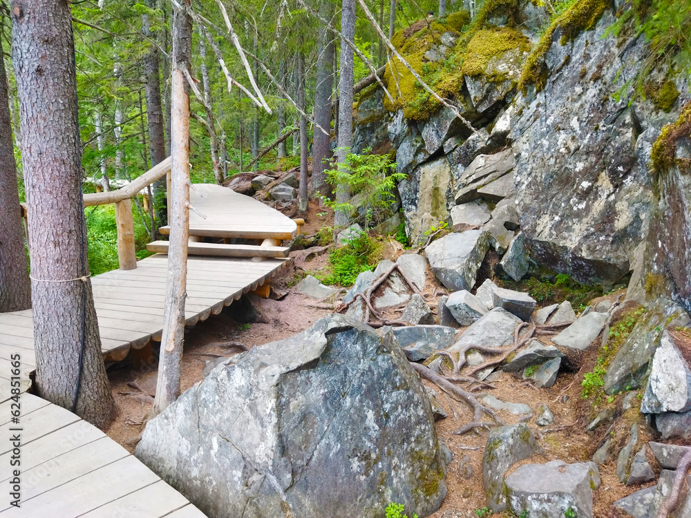 Obraz premium Wooden tourist walking trail as boardwalk in the forest park near Tohmajoki River. Summer water landscape with walkway near Ahvenkoski waterfall, Karelia, Russia.
