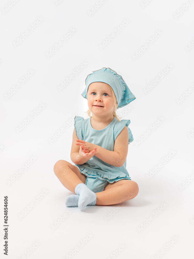 A 2-year-old toddler girl in a kerchief looks up attentively, sits cross-legged on the floor on a white background. A 2-year-old toddler girl in a kerchief looks up attentively, sits cross-legged on the floor on a white background.