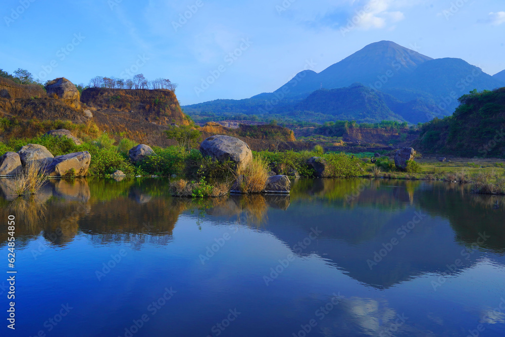A serene pond reflects the surrounding landscape of lush grass, scattered rocks, and towering trees by the breathtaking view of mount Penanggunan in Ranu Manduro, Mojokerto, East Java, Indonesia.