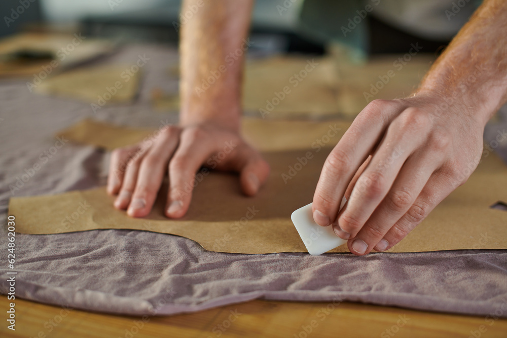 Cropped view of young craftsman marking fabric with chalk while working with sewing pattern on table in print studio, multitasking business owner managing multiple project Cropped view of young craftsman marking fabric with chalk while working with sewing pattern on table in print studio, multitasking business owner managing multiple project