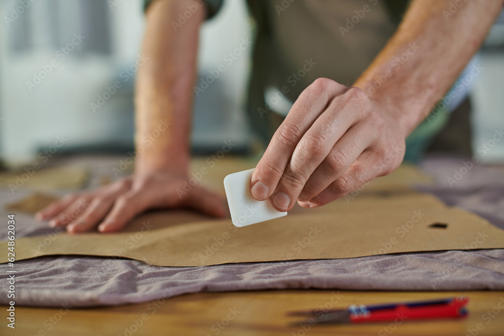 Cropped view of blurred artisan holding crayon near fabric with sewing pattern on table in print studio at background, multitasking business owner managing multiple project