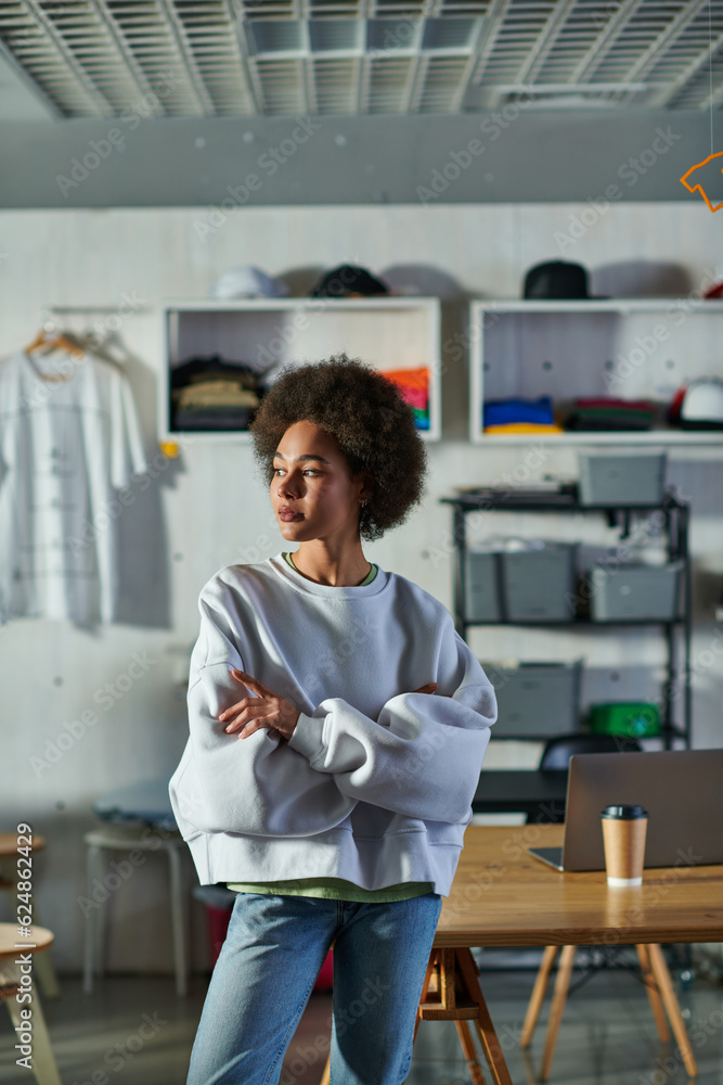Confident young african american craftswoman in sweatshirt and jeans crossing arms while standing in blurred print studio, enthusiastic business owner working in workshop Confident young african american craftswoman in sweatshirt and jeans crossing arms while standing in blurred print studio, enthusiastic business owner working in workshop