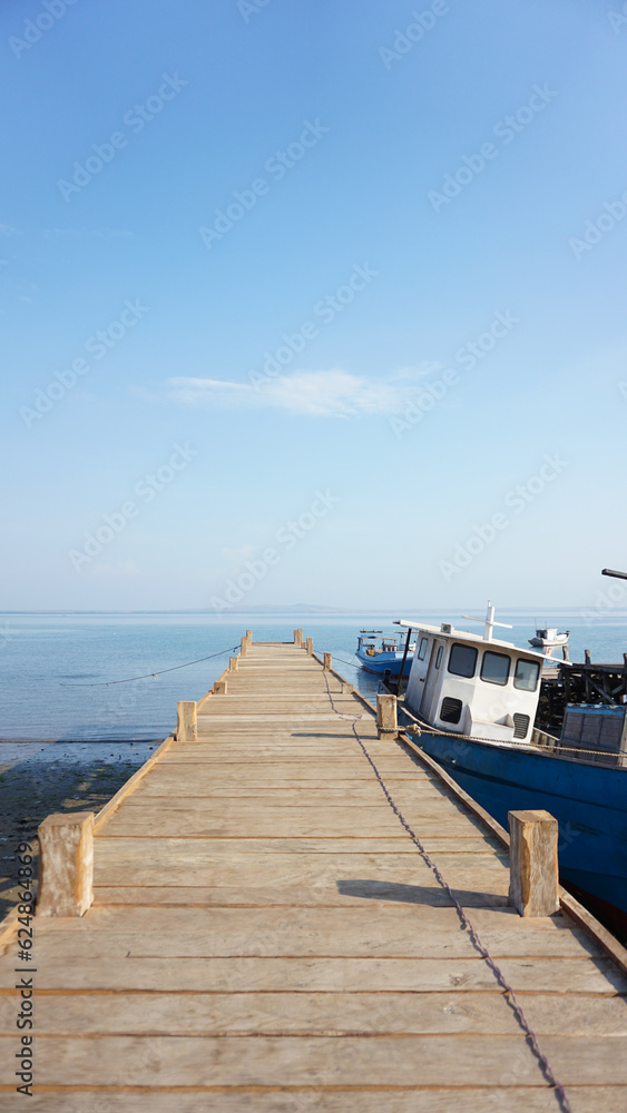 Naklejka premium A wooden bridge Harbor with boats anchored on the island of East Nusa Tenggara. wooden pier bridge with sea and beautiful blue sky. Vertical view. Sea background
