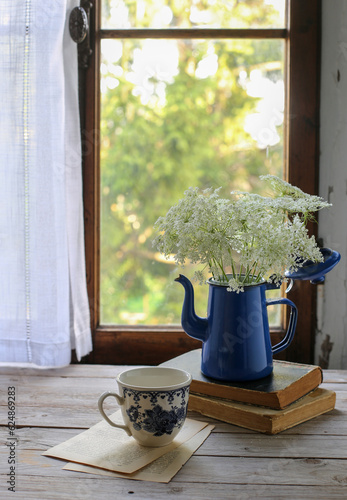 Brocca blu di latta con fiori di carota selvatica sopra alcuni libri di fronte una finestra di legno