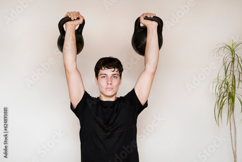 Handsome and strong young teenager guy lifts heavy kettlebells at home, trains his body for strength and endurance, prepares for army service photo