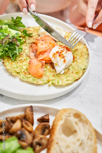 Woman cuts Zucchini pancakes, with avocado, cheese cream, salmon and egg . Healthy breakfast, protein. Restaurant dish. Breakfast in cafe on a sunny morning, on the summer veranda