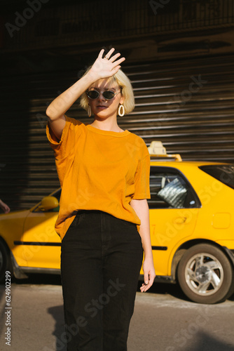 Portrait of young beautiful blonde girl with short hair cut standing on the street near taxi in casual clothes, jeans, t-shirt. Sunny portrait, street style, urban portrait © Ihor