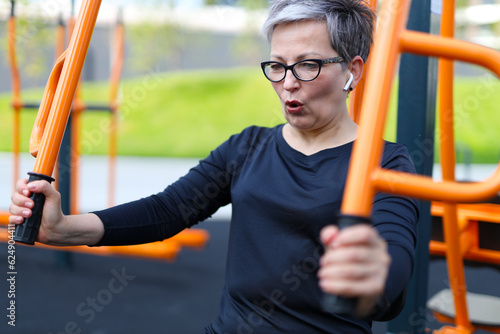A senior woman in sportswear in black clothes and headphones is engaged in an outdoor exercise machine outdoors.