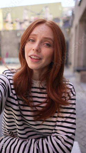 Vertical portrait of cheerful young woman talking video call looking at camera sitting at table in outdoor cafe terrace in sunny day. POV of happy redhead female having video chat using smartphone