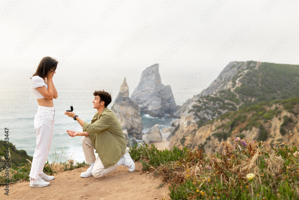 Love on the littoral. Man standing on knee and proposing to the girlfriend with breathtaking coastline view, free space