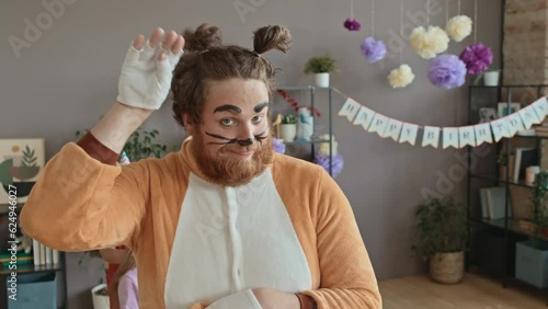 Medium close-up portrait shot of young playful Caucasian male entertainer in cat character costume and makeup performing at kids birthday party, licking its paw while looking at camera