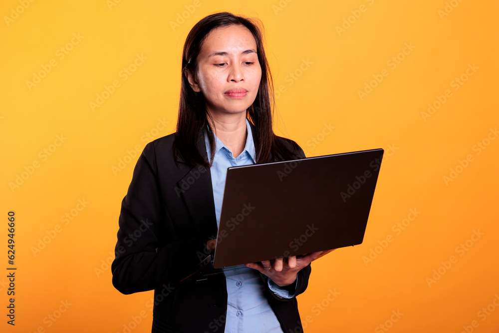 Asian woman holding laptop computer browsing on website, working at research project in studio. Positive model searching information on internet, using device to scroll through social media app