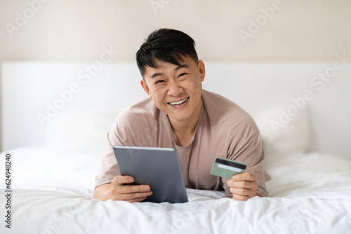 Asian man lying on bed, using tablet and bank card