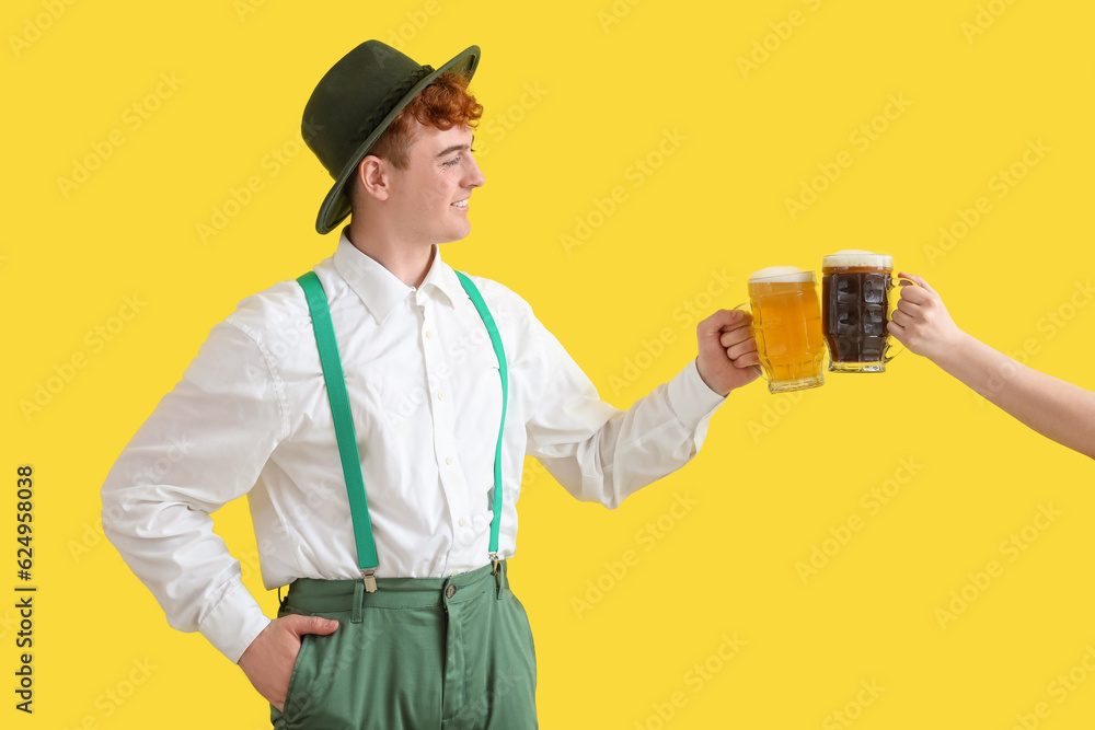 Young man in traditional German clothes and female hand with beer on yellow background