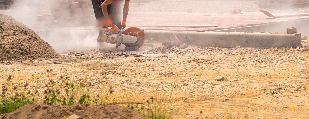 Fototapeta premium Cięcie betonowej kostki brukowej za pomocą piły spalinowej ze stalowa tarczą z diamentami . Chmura pyłu betonowego . Budowa drogi .