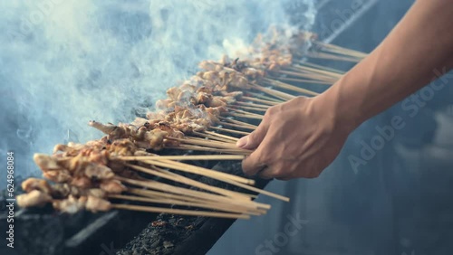  A hand is flipping chicken satay taichan being grilled on a charcoal satay grill while being fanned. The smoke released is quite a lot and thick, spreading everywhere.  photo